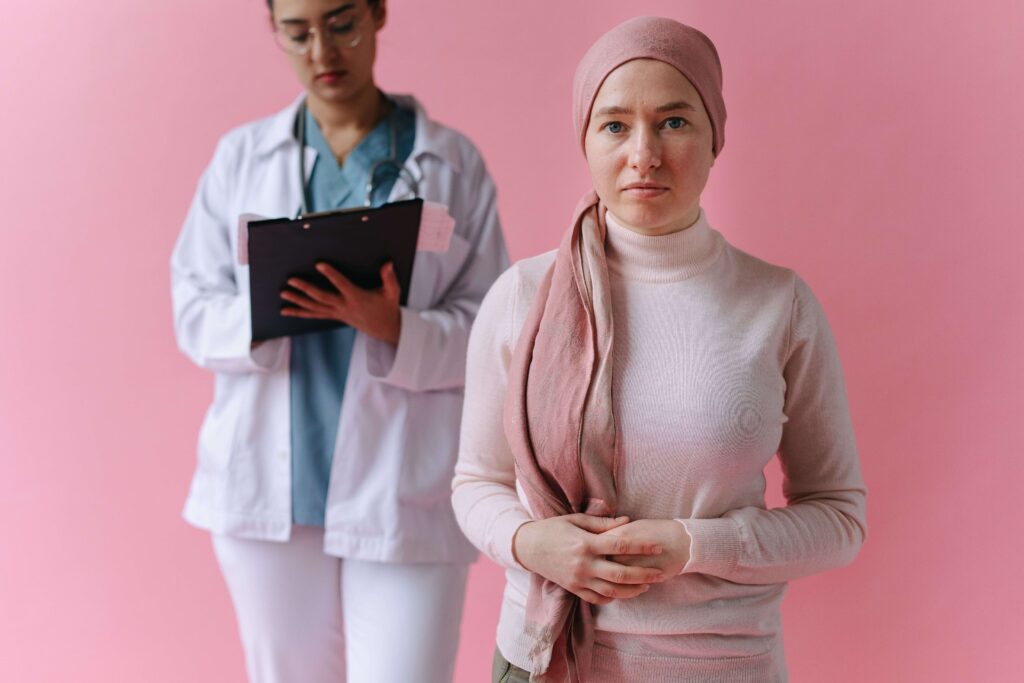 Duas mulheres estão lado a lado. Uma é médica e está segurando uma prancheta e a outra é uma paciente com câncer de mama com blusa cor de rosa e um lenço na cabeça da mesma cor